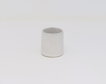 May include: A small, off-white ceramic cup with a cylindrical form. The cup has a slightly textured surface and a smooth, rounded rim. The interior of the cup is a lighter shade of grey. Suitable for holding small items or as a decorative piece.