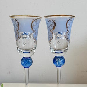 May include: Two elegant champagne flutes. Each glass features a light blue upper section with floral and gold filigree designs. The stem includes a blue orb and a clear, slender base. The rims are trimmed with gold.