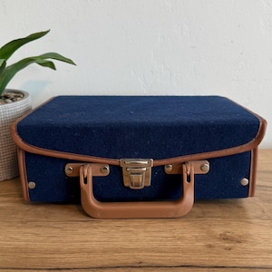 May include: A small, navy blue felt suitcase with a brown leather trim and handle. The suitcase has a silver clasp and is sitting on a wooden surface. A potted plant is visible in the background.