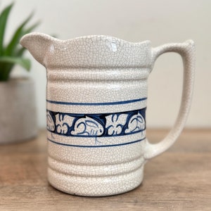 May include: Cream-colored ceramic pitcher with a blue decorative band. The pitcher has a crackle glaze, a spout, and a handle. The blue band features a repeating floral design. A small potted plant is in the background.