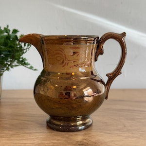May include: A brown ceramic pitcher with a rounded body, a spout, and a curved handle. The pitcher has a glossy finish with decorative floral patterns near the top. The pitcher is sitting on a wooden surface.