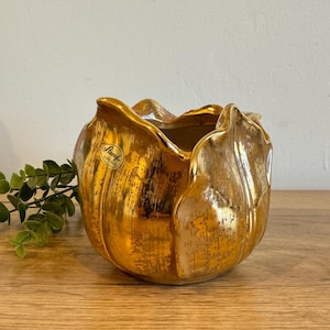 May include: A gold-colored ceramic tulip-shaped vase with a textured finish. The vase has a wide opening and a label on the side. A sprig of green leaves is next to the vase.