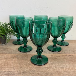 May include: Six emerald green wine glasses with a classic design. The glasses have a stemmed base and a detailed, textured bowl. The glasses are arranged on a wooden surface, with a small potted plant visible in the background.