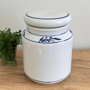 May include: A white ceramic jar with a lid. The jar has a cylindrical shape with a blue floral design and blue trim. The lid is white with a blue trim. The jar is sitting on a wooden surface.
