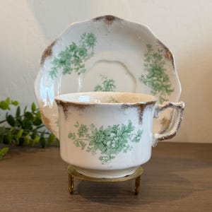 May include: A vintage teacup and saucer set. The white porcelain cup and saucer feature a green floral pattern and gold trim. The cup sits on a small gold-colored stand. The saucer is scalloped.