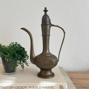 May include: An antique brass pitcher with a long spout and a handle. The pitcher has intricate etched designs and a hinged lid. It is displayed on a stack of books next to a small potted plant.