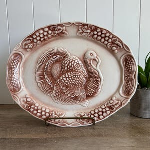 May include: An oval ceramic serving platter featuring a raised, embossed turkey design in the center. The rim is decorated with relief patterns of grapes, leaves, and corn cobs. The platter has a cream-colored base with brown-toned detailing.