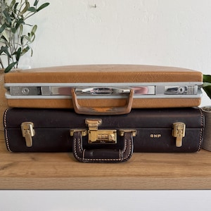 May include: Two vintage briefcases stacked on a wooden surface. The top briefcase is tan with silver hardware and a brown handle. The bottom briefcase is dark brown with gold hardware and a combination lock. The initials "GNP" are visible.