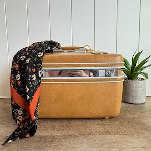 May include: A mustard yellow hard-shell train case with a chrome center band, a top handle, and a key lock. A black scarf with a floral pattern in white, orange, and green is draped over the left side of the rectangular travel vanity case.