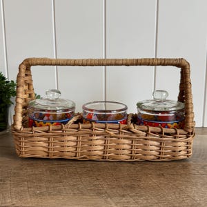 May include: A rectangular woven wicker caddy with a tall arched handle containing three clear glass jars. Each jar features a colorful decorative band with red, yellow, and blue patterns and a matching clear glass lid with a small knob handle.
