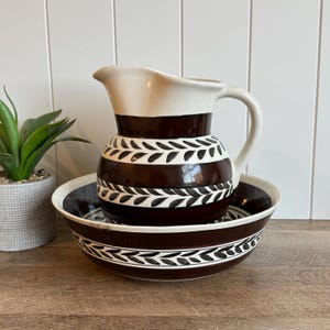 May include: A vintage brown and white ceramic pitcher and bowl set. The pitcher has a white top and handle, with a brown band and leaf design. The bowl has a matching design. The set is on a wooden surface.