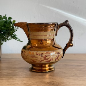 May include: A vintage ceramic pitcher with a metallic gold finish. The pitcher features a bulbous body, a curved handle, and a spout. The design includes horizontal bands and floral patterns in shades of gold and brown. The pitcher sits on a small, round base.
