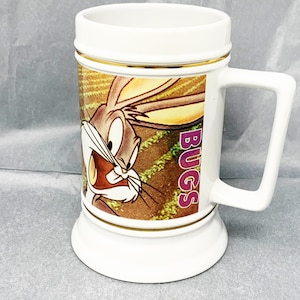 May include: White ceramic Bugs Bunny mug with a gold rim. The mug features a cartoon image of Bugs Bunny with the word "BUGS" printed in purple on the side.