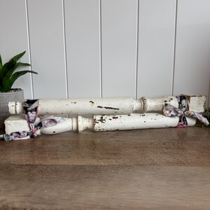 May include: Two white wooden furniture legs with a distressed, chipped paint finish. The legs are cylindrical with square ends and are tied together at both sides with a dark fabric ribbon featuring a floral pattern in shades of pink and purple.