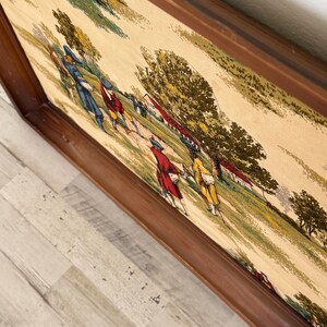 May include: A framed tapestry featuring a pastoral scene with figures in period clothing. The artwork depicts people in red, blue, and yellow coats, walking on a green field with trees and buildings. The frame is made of dark brown wood.