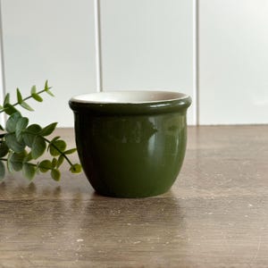 May include: A small, round, olive green ceramic bowl with a smooth, glossy finish and a white interior. The bowl features a slightly flared rim and a deep, rounded base, providing a simple and functional design for various household uses.