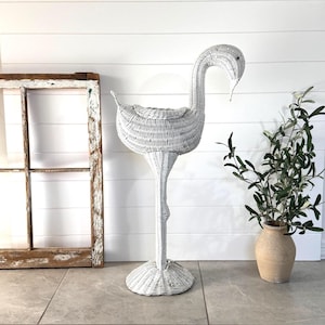 May include: A white wicker sculpture shaped like a long-necked bird, such as a swan or goose, standing on a circular base. The woven structure features a hollow, basket-like body with a curved neck and a pointed beak, crafted from white material.