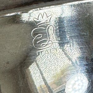 Op de afbeelding: Close-up van een zilverkleurig object met een getextureerd oppervlak. Het object heeft een kroonontwerp boven gestileerde initialen en het woord "ITALY" in hoofdletters. De afbeelding is enigszins onscherp.
