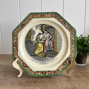 May include: An octagonal ceramic plate featuring a central illustration of two figures in historical clothing, with the text Cries of London printed below. The border is decorated with a brown and tan scrollwork pattern and a green outer rim.