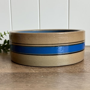 May include: A round, low-profile ceramic bowl with a smooth, tan-colored exterior. The bowl features a prominent, wide horizontal band of vibrant blue glaze encircling the center, bordered by thin, dark brown lines above and below the blue stripe.