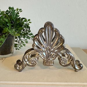 May include: Silver-colored metal napkin holder with an ornate, symmetrical design. The holder features a central, fan-shaped element and decorative scrollwork. The base has a curved shape with floral accents. The holder is placed on a light-colored surface.