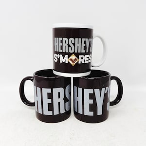 May include: Three brown ceramic mugs with white lettering. The mugs feature the Hershey's S'mores logo. The mugs are stacked on top of each other.