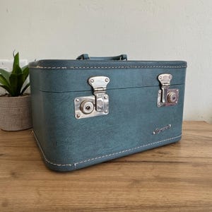 May include: Teal vintage suitcase with silver metal latches and a handle. The rectangular case has white stitching around the edges and a small silver nameplate. The suitcase is sitting on a wooden surface.