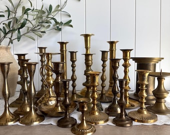 May include: A collection of various brass candlesticks of different heights and shapes, featuring smooth, tapered, and ribbed designs. The metallic candle holders are arranged in a group, displaying a polished gold-toned finish and varied bases.