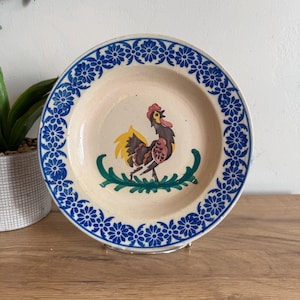 May include: A ceramic plate with a rooster design. The plate has a cream-colored center with a rooster illustration in the center. The rim is decorated with blue floral patterns. The rooster is brown, yellow, and red, standing on a green leafy design.