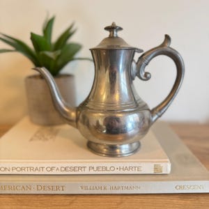 May include: A silver-colored antique teapot with a curved handle and a lid, resting on top of two books. A small potted plant is visible in the background. The teapot has a classic design and is likely made of metal.