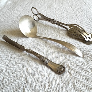 May include: Three antique silver serving utensils: a ladle, a cake server, and a pair of tongs. The ladle has a large, round bowl and a long handle. The cake server has a wide, flat blade. The tongs have a decorative design.