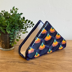 May include: A ceramic napkin holder with a dark blue exterior and a white interior. The holder is decorated with a scale pattern in shades of orange, yellow, and green. The holder is triangular in shape.
