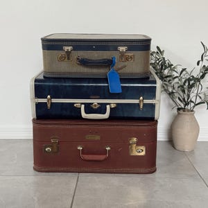 May include: Three vintage suitcases stacked on top of each other. The top suitcase is tan and navy blue, the middle one is navy blue and white, and the bottom one is brown. Each suitcase has metal hardware and a handle. A blue tag hangs from the middle suitcase.