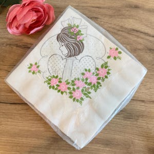 May include: A package of white paper napkins with a vintage illustration of a woman with brown hair and pink roses. The napkins are wrapped in clear plastic and feature a pink rose on the top left.