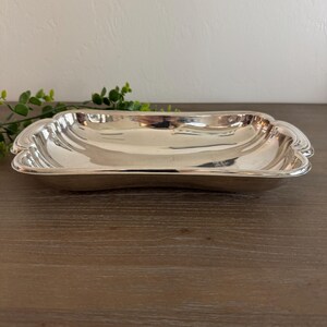 Op de afbeelding: Een rechthoekige, zilverkleurige serveerschaal met een geschulpte rand. De schaal heeft een licht gebogen vorm en een reflecterend oppervlak. De schaal is leeg en staat op een houten oppervlak.