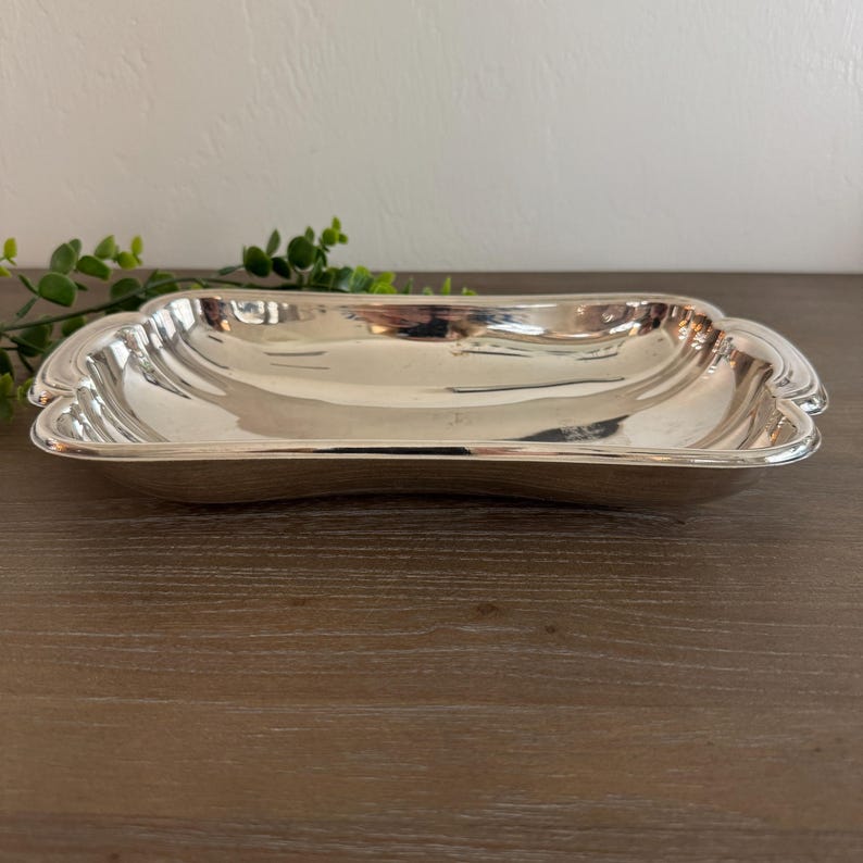Op de afbeelding: Een rechthoekige, zilverkleurige serveerschaal met een geschulpte rand. De schaal heeft een licht gebogen vorm en een reflecterend oppervlak. De schaal is leeg en staat op een houten oppervlak.