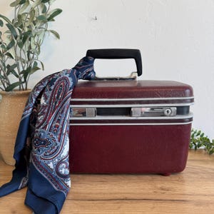 May include: A vintage burgundy vanity case with silver trim and a black handle. A navy blue paisley scarf is draped over the top. The case is on a wooden surface.