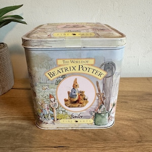 May include: A rectangular tin featuring illustrations from "The World of Beatrix Potter." The tin has a light blue background with images of Peter Rabbit and other characters. The lid has a circular design with the words "Fine Arts."