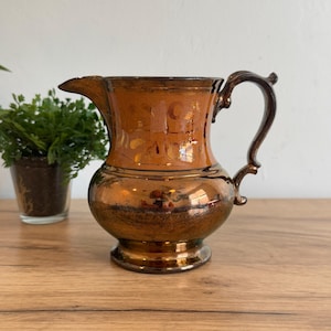 May include: A vintage copper-toned pitcher with a curved handle and spout. The pitcher has a bulbous body and a decorative rim. The surface reflects light, and the handle is a darker shade of brown. A small plant in a glass pot is in the background.