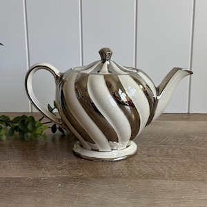 May include: A teapot featuring a swirling pattern of alternating glossy white and reflective silver metallic stripes. The teapot has a curved handle, a pointed spout, and a matching lid with a small knob handle, all finished in a polished metallic sheen.