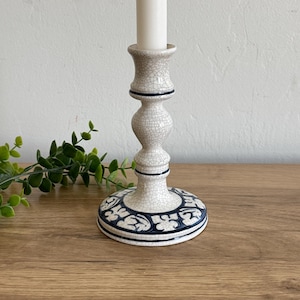 May include: A cream-colored ceramic candlestick with a crackle glaze finish. The base features a blue and white floral design. A white candle sits atop the candlestick. The candlestick is on a wooden surface.