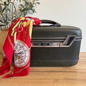 May include: A vintage dark green Amcrest vanity case with a silver handle and accents. A red scarf with a gold watch design is draped over the case. The case is on a wooden surface.