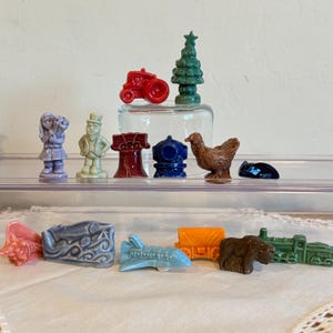 May include: A collection of small, colorful ceramic figurines. The figurines include a red tractor, a green Christmas tree, a blue airplane, a brown bear, and a green train. Other figurines include a girl, a man, a red structure, a blue fire hydrant, and a brown chicken.