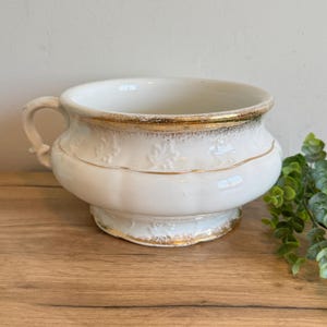May include: A white ceramic bowl with a gold rim and accents. The bowl has a handle and sits on a pedestal base. The bowl has a floral design and is likely vintage or antique.