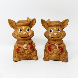 May include: Two brown ceramic bunny figurines with blue eyes and red and white accents. The bunnies are wearing hats and holding a small, round object in their paws.