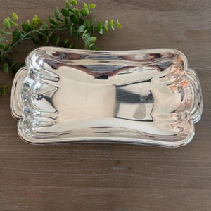 Op de afbeelding: Een rechthoekige, zilverkleurige serveerschaal met een decoratieve, geschulpte rand. De schaal heeft een gebogen vorm en een reflecterend oppervlak, dat de omgeving weerspiegelt. Hij is leeg en ligt op een houten ondergrond.