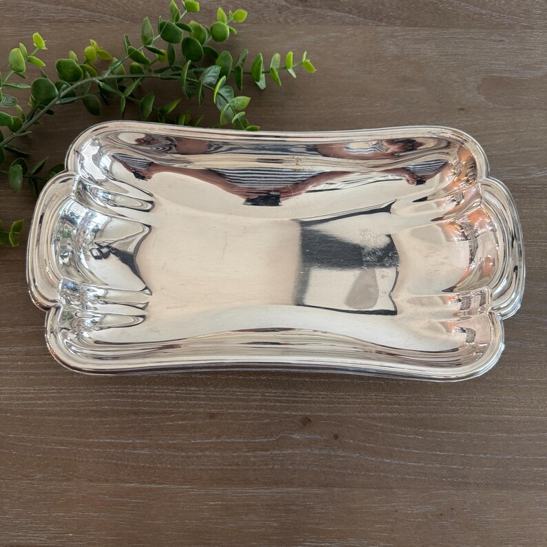 Op de afbeelding: Een rechthoekige, zilverkleurige serveerschaal met een decoratieve, geschulpte rand. De schaal heeft een gebogen vorm en een reflecterend oppervlak, dat de omgeving weerspiegelt. Hij is leeg en ligt op een houten ondergrond.