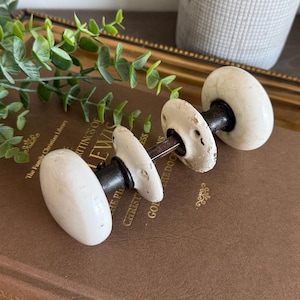 May include: A vintage door knob set featuring two round, off-white ceramic knobs with a subtle crackled finish. The knobs are connected by a dark metal spindle and include two matching circular metal mounting plates with visible screw holes.
