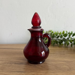 May include: A small, ruby red glass cruet with a handle and a stopper. The cruet has a rounded body and a spout. The stopper is a teardrop shape. The cruet is sitting on a wooden surface.