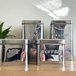 May include: Set of four vintage metal kitchen canisters. The canisters are rectangular with a shiny, reflective surface. Each canister is labeled with embossed lettering: "TEA", "SUGAR", "COFFEE", and "FLOUR". The canisters are arranged on a wooden surface.